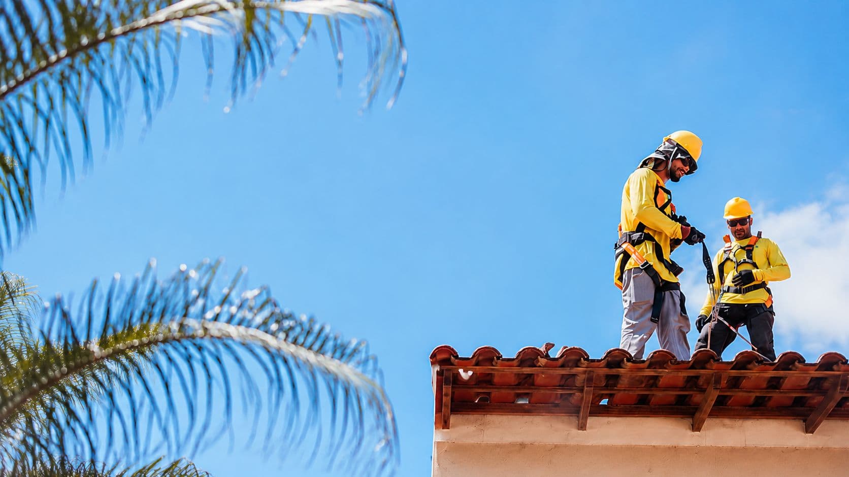 Two roofers in harnesses working along the ridge of a barrel-tile roof under a clear sky